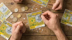 Mani che grattano un biglietto gratta e vinci su un tavolo di legno, con monete e frammenti di pellicola argentata
