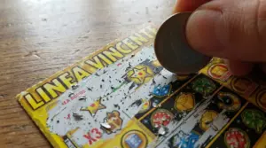 Mano che gratta un biglietto gratta e vinci con una moneta, con simboli del gioco visibili sul tavolo