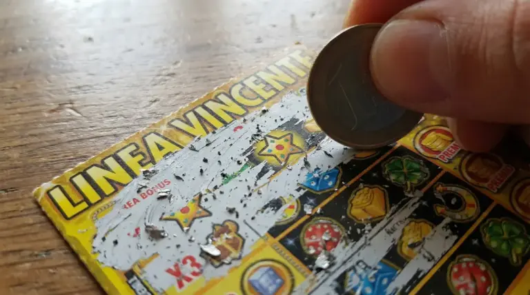 Mano che gratta un biglietto gratta e vinci con una moneta, con simboli del gioco visibili sul tavolo