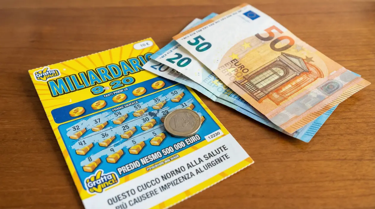 Gratta e vinci su un tavolo con una moneta e banconote in euro accanto, tema vincita e tasse