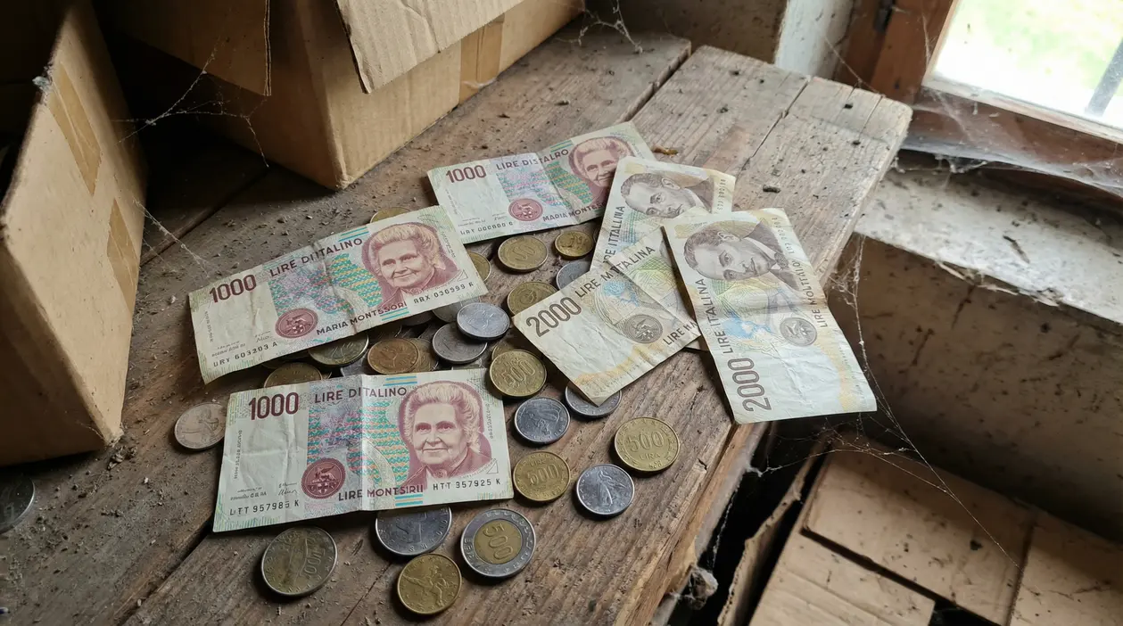 Banconote e monete in lire italiane trovate in soffitta su un tavolo di legno polveroso, vicino a scatole di cartone