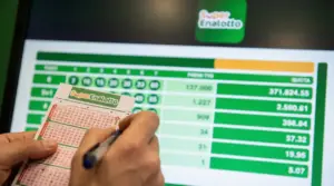 Mani che compilano una schedina del SuperEnalotto davanti a uno schermo con risultati e quote di vincita