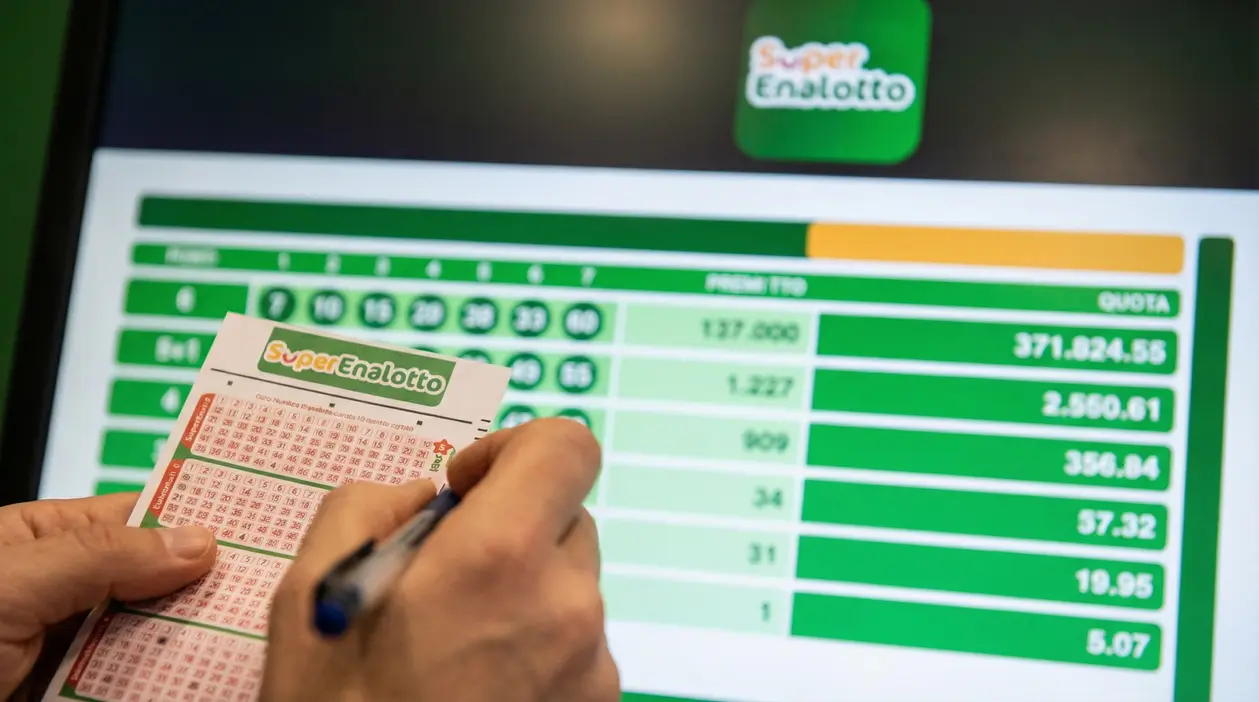 Mani che compilano una schedina del SuperEnalotto davanti a uno schermo con risultati e quote di vincita
